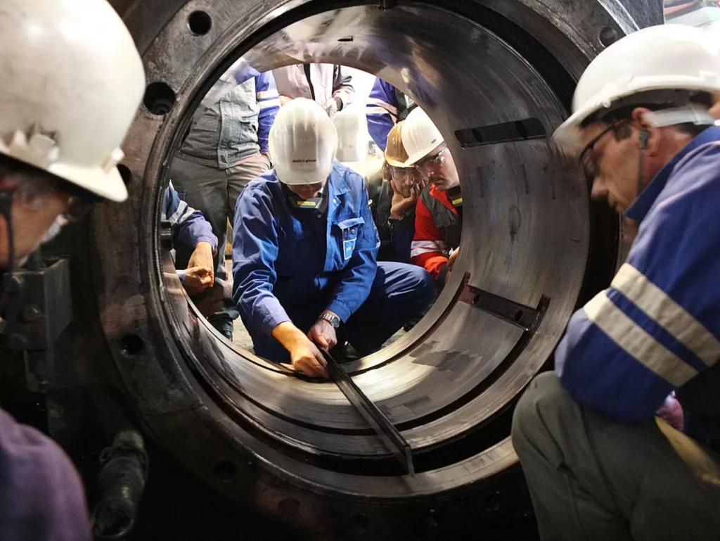 A team of engineers wearing safety helmets and work uniforms collaboratively inspecting a large industrial rolling mill bearing.